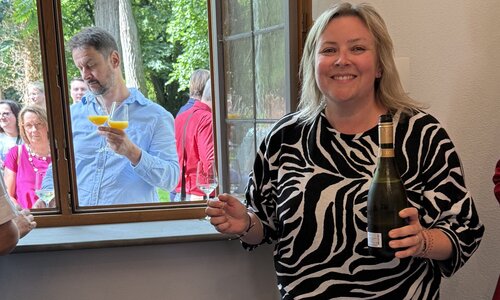 Eine Frau in schwarz-weißem Zebramuster-Oberteil schenkt lächelnd Sekt oder Orangensaft ein. Durch das geöffnete Fenster reicht ein Mann von draußen zwei Gläser herein. Im Hintergrund stehen weitere Gäste.