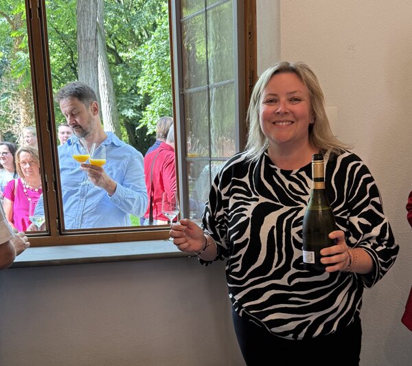 Eine Frau in schwarz-weißem Zebramuster-Oberteil schenkt lächelnd Sekt oder Orangensaft ein. Durch das geöffnete Fenster reicht ein Mann von draußen zwei Gläser herein. Im Hintergrund stehen weitere Gäste.