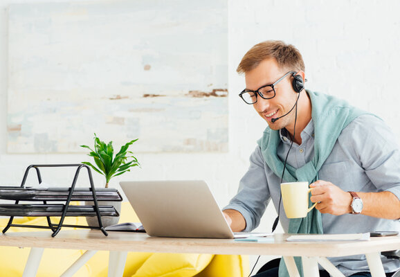 Mann mit Headset arbeitet am Laptop und trinkt Kaffee im Homeoffice
