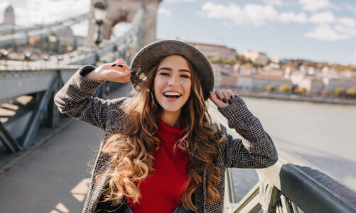 Lächelnde junge Frau mit Hut und kariertem Mantel posiert fröhlich auf einer Brücke in der Stadt bei sonnigem Wetter.