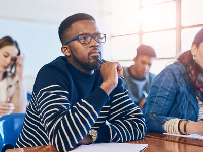 Ein Mann mit Brille und Bart sitzt nachdenklich an einem Tisch in einem Seminar- oder Schulungsraum. Er trägt ein dunkelblau-weiß gestreiftes Langarmshirt und hält einen Stift an seinen Mund, während er konzentriert nach vorne blickt. Im Hintergrund sind weitere Personen unscharf zu erkennen, die ebenfalls an Tischen sitzen und Notizen machen oder zuhören. Der Raum wirkt hell und einladend, mit großen Fenstern, die Tageslicht hereinlassen.