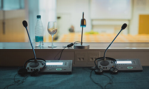 Der Blick zeigt durch die Fensterfront einer Dolmetscherkabine verschwommen in einen Klassenraum. Innerhalb der Kabine stehen zwei identische technische Geräte mit Mikrofon und Kopfhörer, dahinter eine Wasserflasche mit halb gefülltem Glas und eine kleine Lampe.