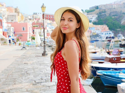 Eine lächelnde Frau mit langen, blonden Haaren steht an einem Hafen oder einer Uferpromenade. Sie trägt ein rotes Kleid mit weißen Punkten und einen Strohhut. Im Hintergrund sind bunte Häuser, Boote im Wasser und eine malerische Küstenlandschaft zu sehen. Die Frau wirkt entspannt und genießt den sonnigen Tag. Der Himmel ist klar, und die Atmosphäre wirkt mediterran und einladend.