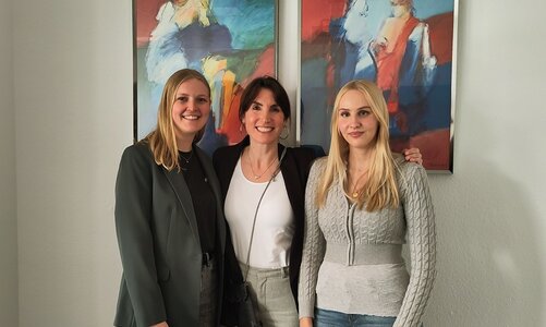 Drei Frauen stehen nebeneinander vor einer weißen Wand in der WDS in Würzburg. Im Hintergrund hängen zwei farbintensive Gemälde mit abstrakten Motiven in Rot-, Blau- und Gelbtönen. Die Frauen tragen unterschiedliche Oberteile: eine schwarze Jacke, ein weißes Oberteil und ein grauer Strickpullover. Alle blicken lächelnd zur Kamera und posieren für ein Erinnerungsfoto.