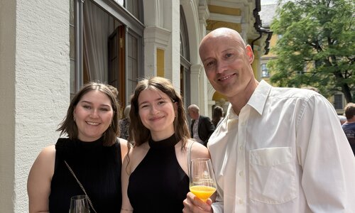 Zwei junge Frauen in schwarzen Kleidern und ein Mann im weißen Hemd stehen draußen vor einem festlichen Gebäude und halten Gläser mit Orangensaft. Sie lächeln in die Kamera.