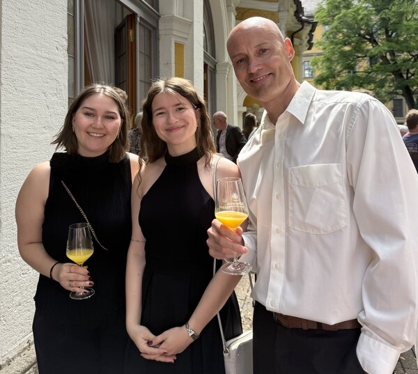 Zwei junge Frauen in schwarzen Kleidern und ein Mann im weißen Hemd stehen draußen vor einem festlichen Gebäude und halten Gläser mit Orangensaft. Sie lächeln in die Kamera.