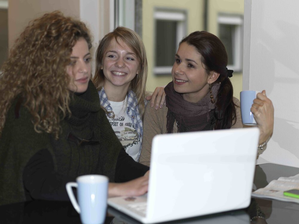 Fröhliche Zusammenarbeit am Laptop Drei Personen sitzen an einem Tisch mit Laptop und blauen Tassen