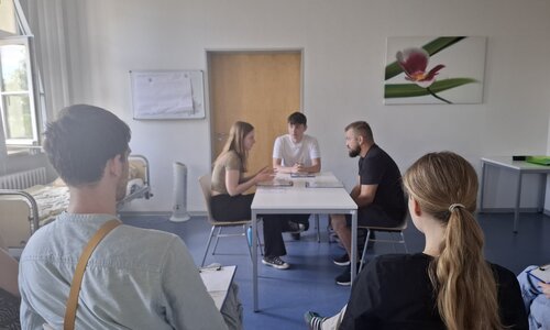 In einem Raum mit weißen Wänden und dunkelblauem Boden sitzen an einem weißen Tisch zwei junge Männer und eine junge Frau. Sie haben Notizen vor sich liegen und unterhalten sich intensiv. Im Vordergrund sitzen Zuschauer, die sich Notizen machen. Am linken Bildrand steht ein leeres Krankenbett.