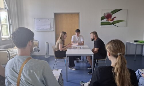 In einem Raum mit weißen Wänden und dunkelblauem Boden sitzen an einem weißen Tisch zwei junge Männer und eine junge Frau. Sie haben Notizen vor sich liegen und unterhalten sich intensiv. Im Vordergrund sitzen Zuschauer, die sich Notizen machen. Am linken Bildrand steht ein leeres Krankenbett.
