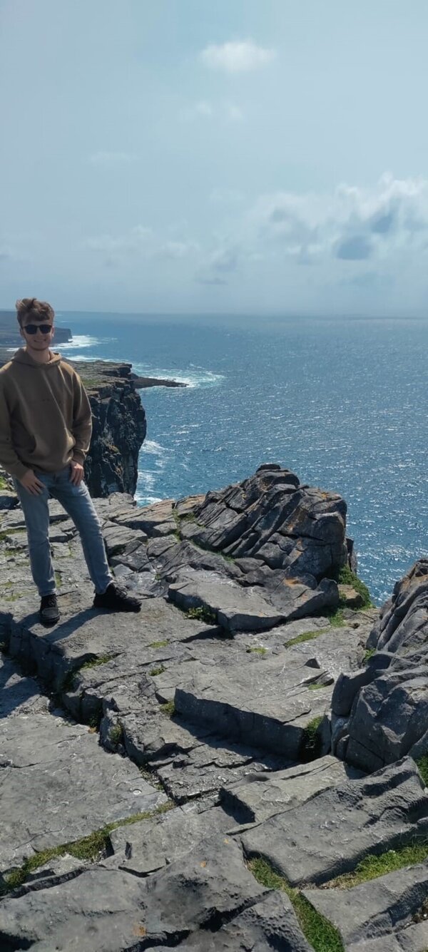Laurin Maiberger an der irischen Küste Der WDS-Schüler Laurin Maiberger hat sich für das Foto an den Rand der Steilküste gestellt. Zwischen ihm und dem Abhang sind nur ein paar Schritte. Der Untergrund ist felsig. Weit unten kann man die Wellen erkennen, die sich an den Klippen brechen. Das Wetter ist sonnige, in der Ferne sieht man ein paar vereinzelte Wölkchen.