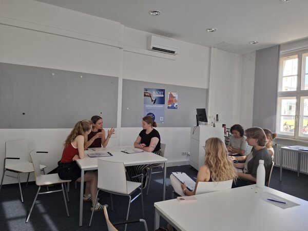 In einem hellen Klassenzimmer sitzen an einem weißen Tisch drei junge Frauen. Sie haben Notizen vor sich liegen und unterhalten sich intensiv. Links vorne sitzen vier weitere Personen, schauen zu und machen sich Notizen.