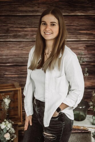 Junge Frau mit langen, glatten Haaren steht lächelnd vor einer Holz­wand, trägt ein weißes Shirt mit offener weißer Bluse, dunkle Jeans mit Riss und eine Armbanduhr, eine Hand lässig in der Hosentasche; im Vordergrund sind dezent Blumen und Deko zu sehen.
