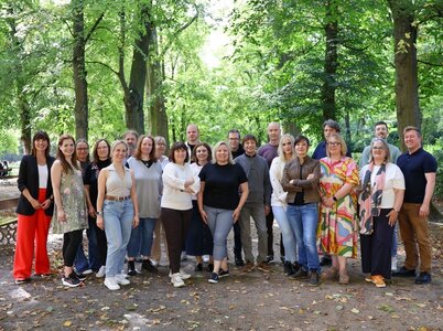 Eine Gruppe von 21 Personen steht in einem Wald oder Park auf einem mit Blättern bedeckten Weg. Die Personen sind in zwei Reihen angeordnet und lächeln in die Kamera. Die Umgebung ist grün und naturnah, mit hohen Bäumen und dichtem Laub im Hintergrund. Die Personen tragen verschiedene Kleidungsstücke, darunter Blusen, Hemden, Pullover, Jeans und Schuhe. Einige haben die Arme verschränkt, andere stehen entspannt mit den Händen an den Seiten.