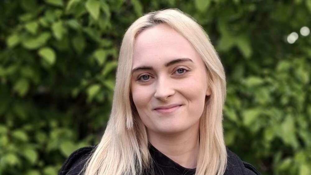 Eine junge Frau mit glatten, langen, blonden Haaren und dunklen Augenbrauen grinst mit verschmitztem Blick in die Kamera. Sie trägt ein schwarzes Oberteil und steht vor eine begrünten Wand.