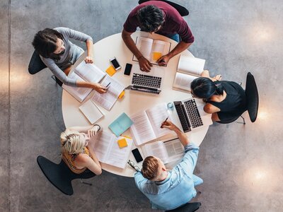 Fünf Personen arbeiten gemeinsam an einem runden Tisch mit Büchern, Laptops und Notizblöcken
