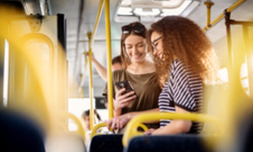 Zwei junge Frauen stehen in einem Bus und schauen lächelnd auf ein Handy. Sie tragen kurze T-Shirt, eine der beiden hat eine Sonnenbrille im Haar. Die Haltestangen des Busses bringen gelbe Farbkleckse ins Bild.