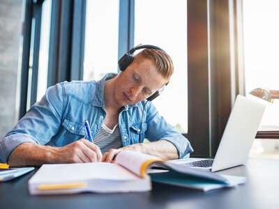 Student beim Lernen Ein junger Student sitzt an einem Schreibtisch direkt am Fenster und macht Notizen. Auf dem Tisch befinden sich ein Laptop und mehrere Bücher. Der Mann trägt ein lässiges Jeanshemd, darunter ein geringeltes Shirt. Er hat große, schwarze Kopfhörer auf.