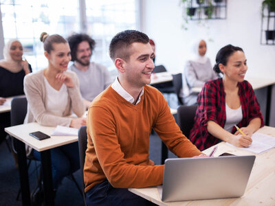 In einem hellen Seminarraum sitzen mehrere Erwachsene an Tischen und nehmen an einer Schulung oder einem Kurs teil. Im Vordergrund sitzt ein lächelnder Mann in einem orangefarbenen Pullover und arbeitet an einem Laptop. Neben ihm sitzt eine lächelnde Frau in einem karierten Hemd, die Notizen macht. Im Hintergrund sind weitere Teilnehmer zu sehen, darunter eine Frau mit Hijab und eine weitere Person, die ebenfalls Notizen macht. Der Raum wirkt modern und einladend, mit großen Fenstern und Pflanzen an den Wänden.
