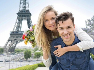 Zwei Personen stehen vor dem Eiffelturm in Paris. Die eine Person hält einen farbenfrohen Blumenstrauß, während die andere sie liebevoll umarmt. Der Himmel ist klar und die Szenerie strahlt Romantik aus.