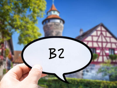 Eine Hand hält eine Sprechblase mit dem Text 'B2'. Im Hintergrund ist eine malerische Szene mit traditionellen europäischen Gebäuden zu sehen, darunter ein Fachwerkhaus und ein Gebäude mit einem turmartigen Aufbau. Im Hintergrund sind auch Bäume und Grünflächen sichtbar.