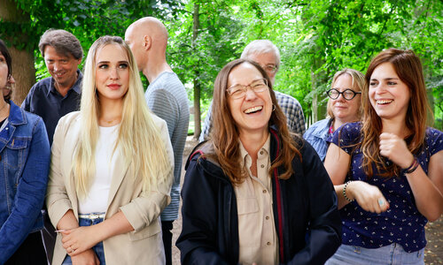 Eine Gruppe lachender Frauen und Männer vor einem Hintergrund aus grünen Bäumen.