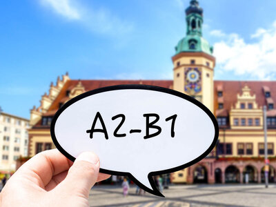 Eine Hand hält eine Sprechblase mit dem Text 'A2-B1'. Im Hintergrund ist ein historisches Gebäude mit einem Uhrturm und architektonischen Details zu sehen, wahrscheinlich in einem europäischen Stadtplatz.