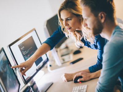 Eine Frau und ein Mann arbeiten zusammen an einem Computer-Arbeitsplatz. Die Frau zeigt auf einen der beiden Bildschirme, auf dem Programmiercode zu sehen ist, während der Mann mit der Maus arbeitet. Beide konzentrieren sich auf die Aufgabe, was eine kooperative und technische Arbeitsumgebung widerspiegelt.