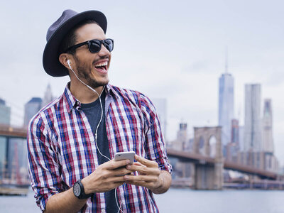 Ein lächelnder Mann mit Sonnenbrille, grauer Kappe und Kopfhörern steht im Freien und hält ein Smartphone in der Hand. Er trägt ein rot-weiß-kariertes Hemd über einem dunklen T-Shirt und wirkt entspannt und fröhlich. Im Hintergrund sind die Silhouetten einer amerikanischen Großstadt mit Hochhäusern und einer berühmten Brücke zu erkennen.