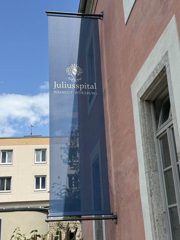 Ein blaues Banner mit der Aufschrift „Juliusspital – Weingut Würzburg“ hängt an einer rosa Außenwand. Der Himmel ist blau mit wenigen Wolken.
