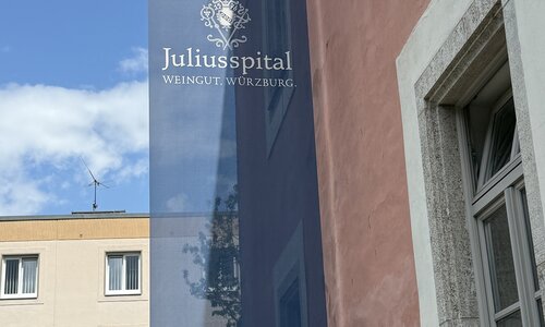 Ein blaues Banner mit der Aufschrift „Juliusspital – Weingut Würzburg“ hängt an einer rosa Außenwand. Der Himmel ist blau mit wenigen Wolken.