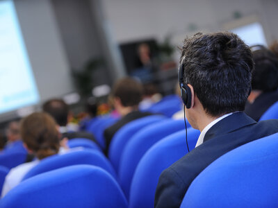 Teilnehmer sitzen auf blauen Stühlen bei einer Konferenz; im Vordergrund ein Mann mit Kopfhörern
