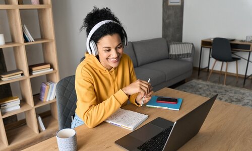 Eine junge Frau mit gelbem Pulli und Kopfhörern sitzt lächelnd vor einem Laptop. Hinter ihr ist ein gemütliches Zimmer zu sehen.