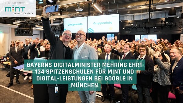 In einem modernen, gut beleuchteten Veranstaltungsraum mit sichtbaren Deckenlampen und technischen Installationen steht eine große Gruppe von Menschen dicht beieinander. Im Vordergrund nehmen zwei Männer ein Selfie mit einem Smartphone, während die übrigen Personen im Hintergrund in die Kamera winken. Auf zwei großen Bildschirmen im Hintergrund ist der Schriftzug 'Gruppenfoto' zu sehen. Im unteren Bildbereich befindet sich ein grüner Balken mit weißer Schrift, der den Text 'BAYERNS DIGITALMINISTER MEHRING EHRT 134 SPITZENSCHULEN FÜR MINT UND DIGITAL-LEISTUNGEN BEI GOOGLE IN MÜNCHEN' zeigt. Links oben ist das Logo der Nationalen Initiative MINT Zukunft schaffen in den Farben Blau und Grün zu erkennen. Die Personen tragen überwiegend formelle Kleidung wie Anzüge und Hemden. Die Szene vermittelt den Eindruck einer feierlichen Auszeichnung im Bereich MINT und Digitalisierung.
