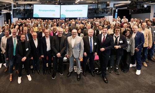 Eine große Gruppe von Menschen steht in einem modernen, gut beleuchteten Veranstaltungsraum. Die Personen sind in mehreren Reihen angeordnet, die vorderen Reihen stehen, während die hinteren Reihen teilweise auf Stühlen sitzen. Die Kleidung der Anwesenden ist überwiegend formell bis business-casual, mit Anzügen, Blazern, Hemden und Blusen in verschiedenen Farben, darunter Grau, Blau, Schwarz und Beige. Im Hintergrund sind zwei große Bildschirme zu sehen, auf denen jeweils das Wort 'Gruppenfoto' in blauer Schrift auf weißem Hintergrund angezeigt wird. Die Decke ist mit mehreren schwarzen Lampen und technischen Geräten ausgestattet. Der Raum wirkt modern und eignet sich für größere Veranstaltungen oder Konferenzen. Das Bild entstand im Rahmen der dritten Auszeichnung der Würzburger Dolmetscherschule zur MINT-freundlichen Schule.