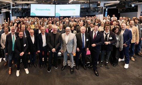 Eine große Gruppe von Menschen steht in einem modernen, gut beleuchteten Veranstaltungsraum. Die Personen sind in mehreren Reihen angeordnet, die vorderen Reihen stehen, während die hinteren Reihen teilweise auf Stühlen sitzen. Die Kleidung der Anwesenden ist überwiegend formell bis business-casual, mit Anzügen, Blazern, Hemden und Blusen in verschiedenen Farben, darunter Grau, Blau, Schwarz und Beige. Im Hintergrund sind zwei große Bildschirme zu sehen, auf denen jeweils das Wort 'Gruppenfoto' in blauer Schrift auf weißem Hintergrund angezeigt wird. Die Decke ist mit mehreren schwarzen Lampen und technischen Geräten ausgestattet. Der Raum wirkt modern und eignet sich für größere Veranstaltungen oder Konferenzen. Das Bild entstand im Rahmen der dritten Auszeichnung der Würzburger Dolmetscherschule zur MINT-freundlichen Schule.