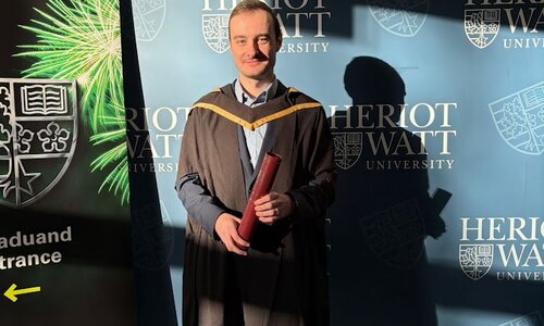 Absolvent steht in einem schwarzen Talar mit gelbem Besatz und hält lächelnd eine rote Urkunde in beiden Händen. Im Hintergrund befindet sich eine dunkelblaue Wand mit dem Schriftzug und Wappen von der Heriot-Watt University. Links ist ein Roll-up mit grünem Feuerwerksmotiv und weißem Text zu sehen.