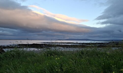 Melancholisches Galway Ein Küste in Irland: Im Vordergrund sieht man Gräser, dahinter einen Küstenstreifen und das Meer, in der Ferne Berge. Die Wolken hängen tief. Die Szenerie wirkt melancholisch. Eine helle Wolke, die von der Sonne angestrahlt wird, lockert die finstere Atmosphäre auf. Ganz links im Bild kann man eine Frau erkennen.