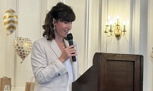 Eine Frau im hellen Anzug steht an einem Rednerpult und spricht in ein Mikrofon. Im Hintergrund befinden sich herzförmige Luftballons in Gold-Weiß und dekorierte Tische.