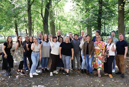 Eine Gruppe von 21 Personen steht auf einem mit Blättern bedeckten Weg in einem Wald oder Park. Die Personen sind in zwei Reihen angeordnet und wirken fröhlich. Einige zeigen Handgesten wie den „Peace“-Zeichen oder Daumen hoch. Die Umgebung ist grün und naturnah, mit hohen Bäumen und dichtem Laub im Hintergrund. Die Personen tragen verschiedene Kleidungsstücke, darunter Blusen, Hemden, Pullover, Jeans und Schuhe. Die Stimmung wirkt locker und freundschaftlich.