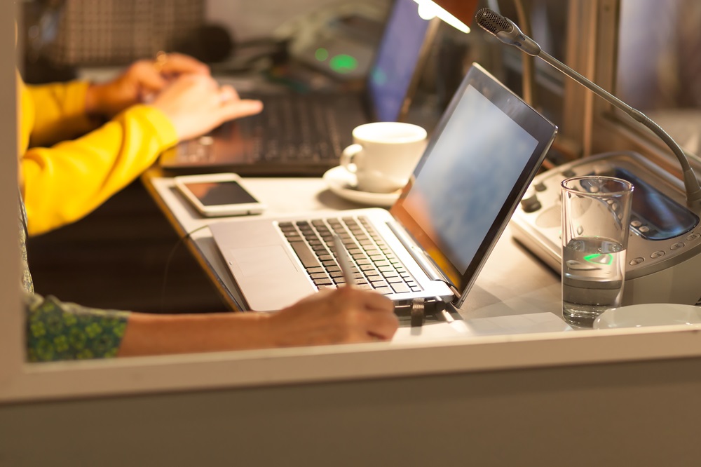 Blick in eine Dolmetscherkabine: Zwei Laptops stehen auf einem Tisch. An einem sieht man Hände, die tippen. Die andere Person macht sich mit einem Stift händische Notizen. Vor dem Laptop ist eine Art Sprechanlage mit Mikrofon, außerdem stehen dort eine Kaffeetasse und ein Glas Wasser.