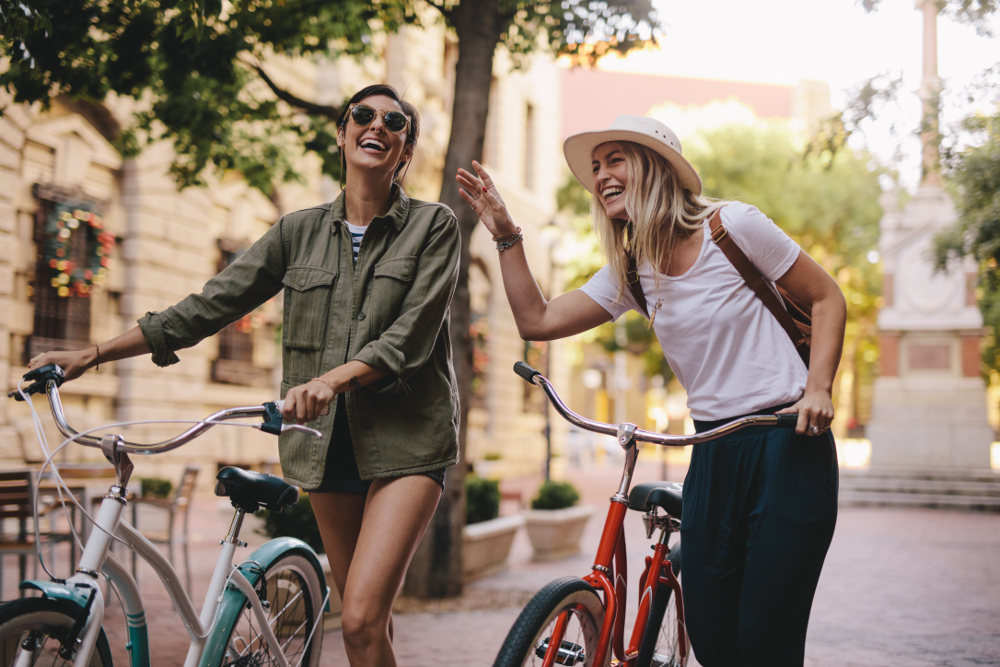 Freundinnen genießen einen fröhlichen Fahrradausflug in der Stadt Zwei Frauen stehen im Freien und lachen, während sie ihre Fahrräder halten. Eine der Frauen trägt eine grüne Jacke und Sonnenbrille, die andere trägt einen weißen Hut und ein weißes T-Shirt. Sie scheinen eine angenehme Zeit in einer urbanen Umgebung mit Bäumen und Gebäuden im Hintergrund zu haben.