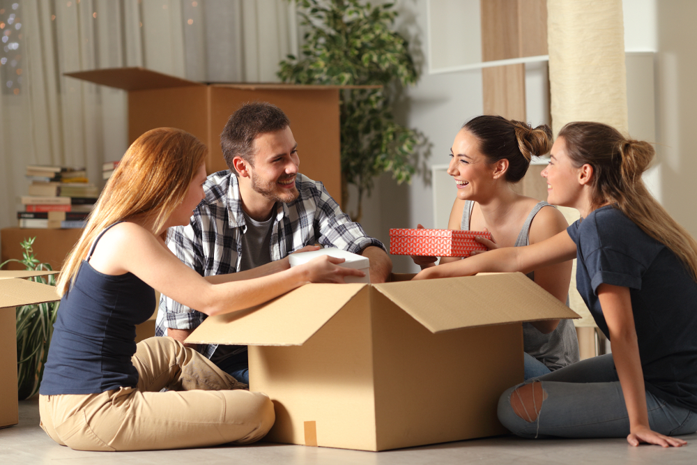 Freude beim gemeinsamen Auspacken eines Kartons Vier Personen sitzen auf dem Boden vor einem geöffneten Karton. Zwei der Personen halten kleine bunte Boxen und scheinen sich über den Inhalt des Kartons zu freuen. Die Szene findet in einem Raum mit Pflanzen und Büchern im Hintergrund statt, was auf eine gemütliche und freundliche Atmosphäre hindeutet.