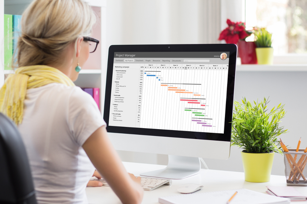 Eine Person sitzt an einem Schreibtisch und betrachtet einen Computerbildschirm, auf dem eine Projektmanagement-Software mit einem Gantt-Diagramm angezeigt wird. Der Schreibtisch ist mit einem kleinen Topfpflanze, einem Stiftebecher und anderen Büroutensilien ausgestattet. Die Person trägt ein weißes T-Shirt und ein gelbes Halstuch.