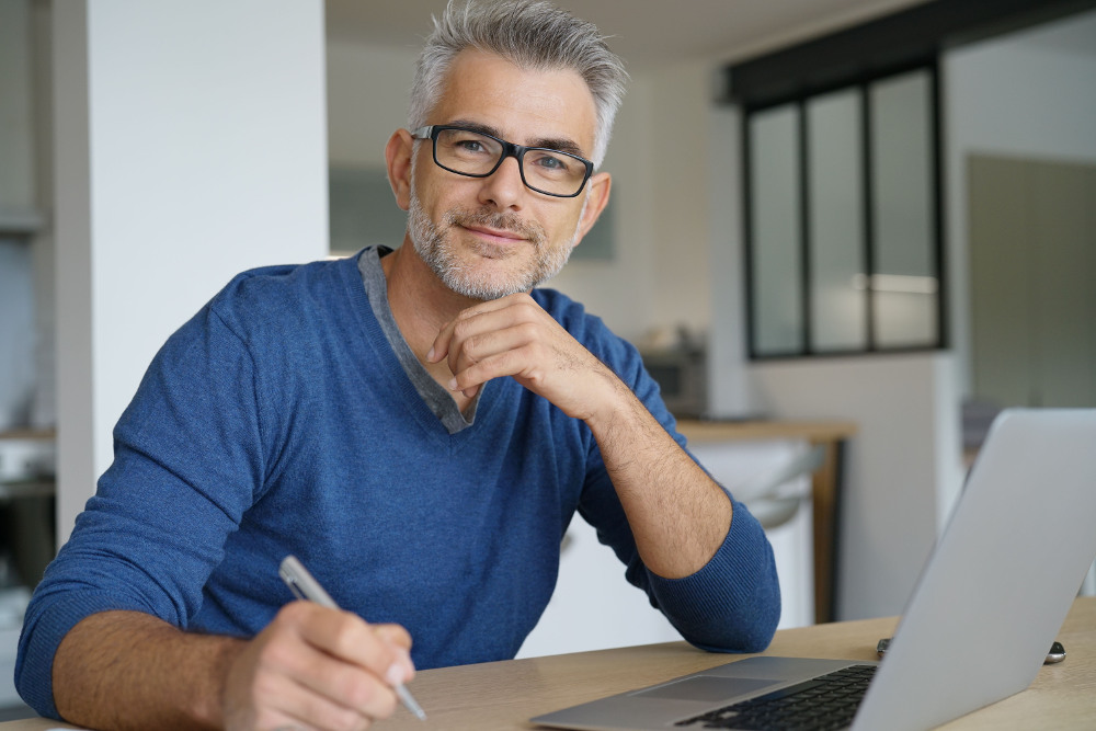 Mann mit Brille arbeitet am Laptop Ein Mann mit blauem Pullover, Brille und grauen Haaren und Bart sitzt an einem Schreibtisch vor einem Laptop. Er schaut leicht lächelnd und nachdenklich in die Kamera. In einer Hand hält er einen Stift.